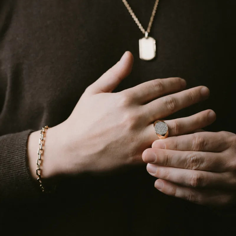 A close-up lifestyle shot of a person's hands wearing jewelry, featuring a rose gold signet ring with a textured bezel, a gold chain necklace with a rectangular tag pendant, and a gold chain bracelet, all set against a dark brown sweater.