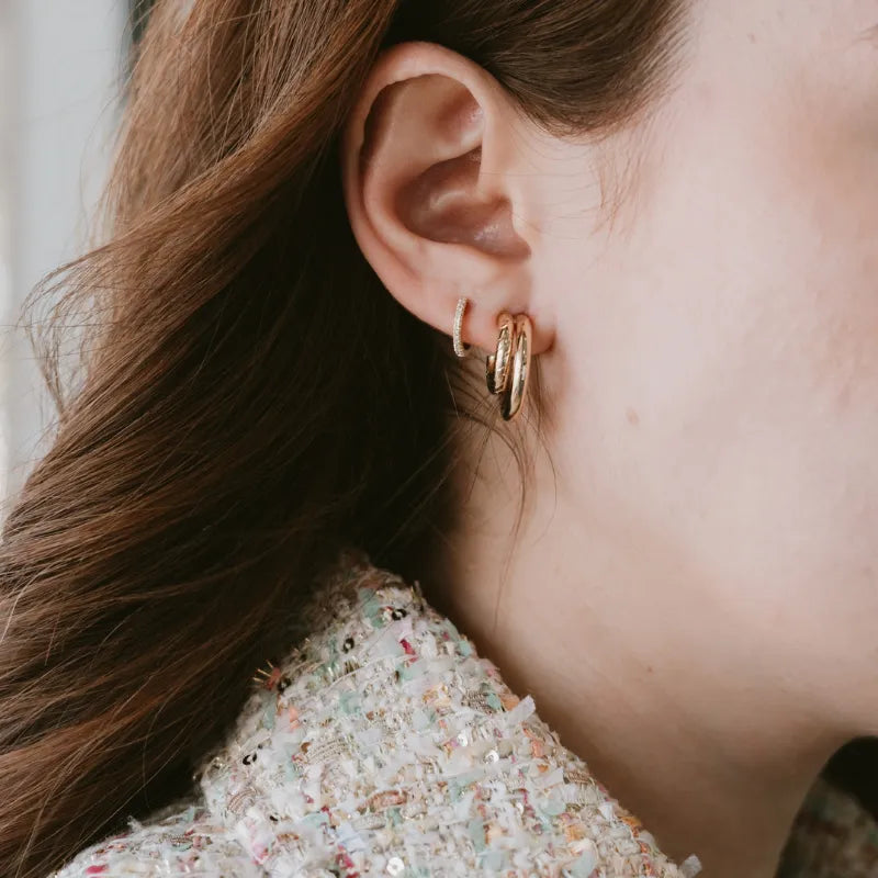 A close-up of a person's ear wearing two small gold hoop earrings, with brown hair and part of a textured tweed top visible.