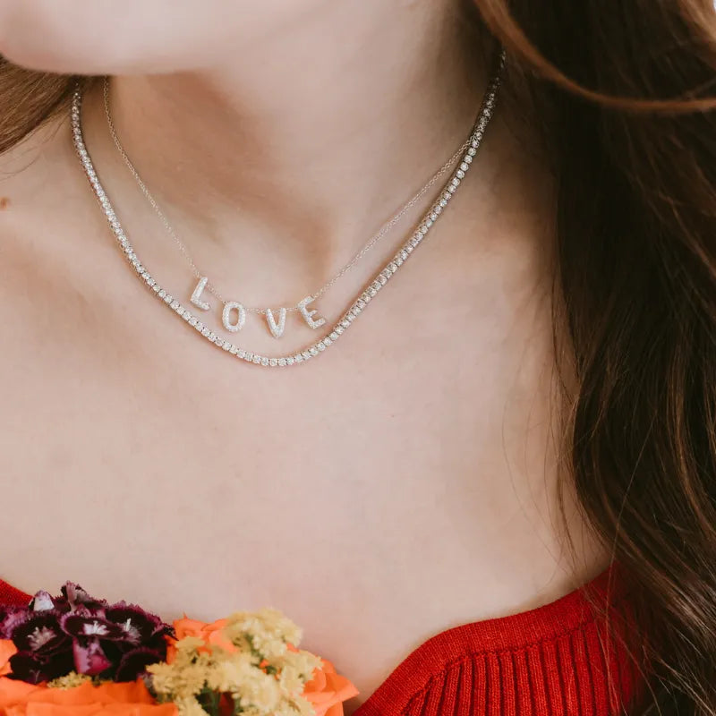 A close-up of a woman wearing a double-layered silver necklace. The top layer is a delicate chain, and the bottom layer features a row of clear rhinestones with the word 'LOVE' spelled out in silver letter charms.