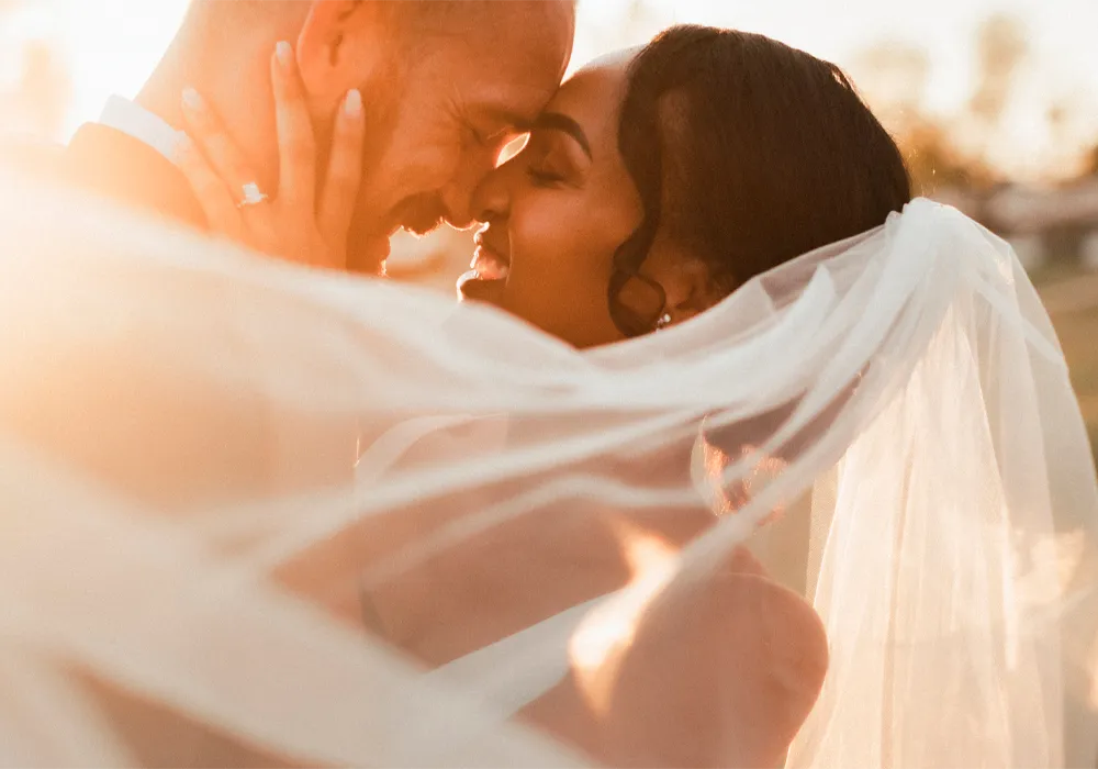 A close-up wedding scene with a bride and groom embracing, featuring a flowing white veil and warm golden sunlight in a softly blurred outdoor setting.
