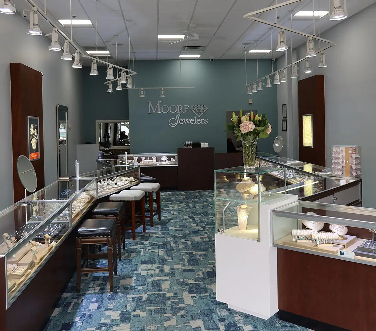 A jewelry store interior with multiple glass display cases, white and wood counters, hanging track lights, and a blue patterned floor. The wall displays the brand name 'Moore's Jewelers'.