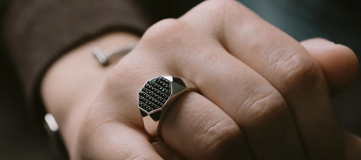 A close-up of a hand wearing a silver-toned signet ring with a textured black inlay on the face of the ring.