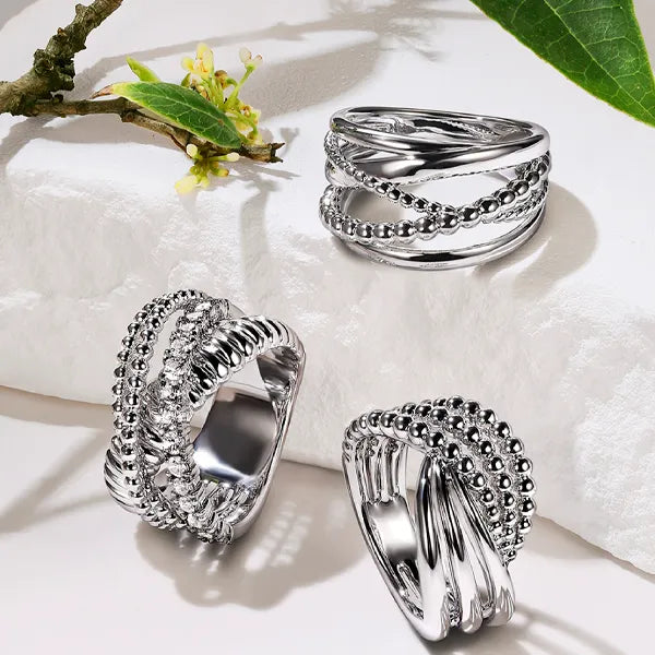 Three silver-toned twisted beaded rings displayed on a white textured surface with a small green leaf branch in the corner.