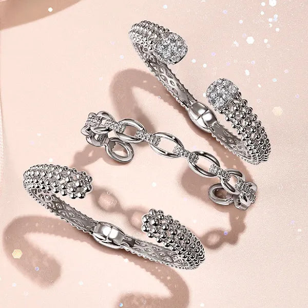 Four silver-toned open cuff bracelets arranged on a soft pink background. Each bracelet features textured pavé-set clear stones and a chain-link segment, with sparkling light effects around them.