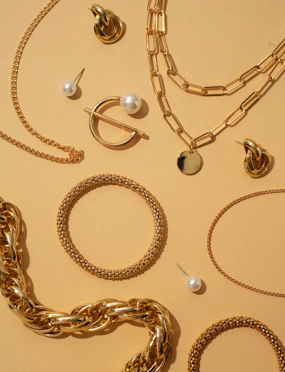 A styled flat-lay of various gold-toned jewelry pieces, including chunky chain necklaces, textured bangles, stud earrings, and a pearl drop earring, arranged on a warm beige background.