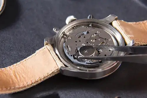 Close-up of a watch back with a transparent case cover showing a detailed mechanical movement, with tweezers holding a component, and a tan leather strap on a dark textured background.