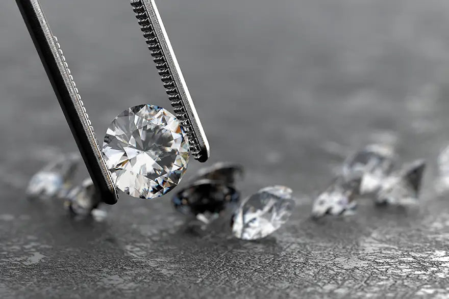 A round brilliant-cut diamond held by silver tweezers against a dark gray textured background with other loose diamonds blurred in the foreground and background.