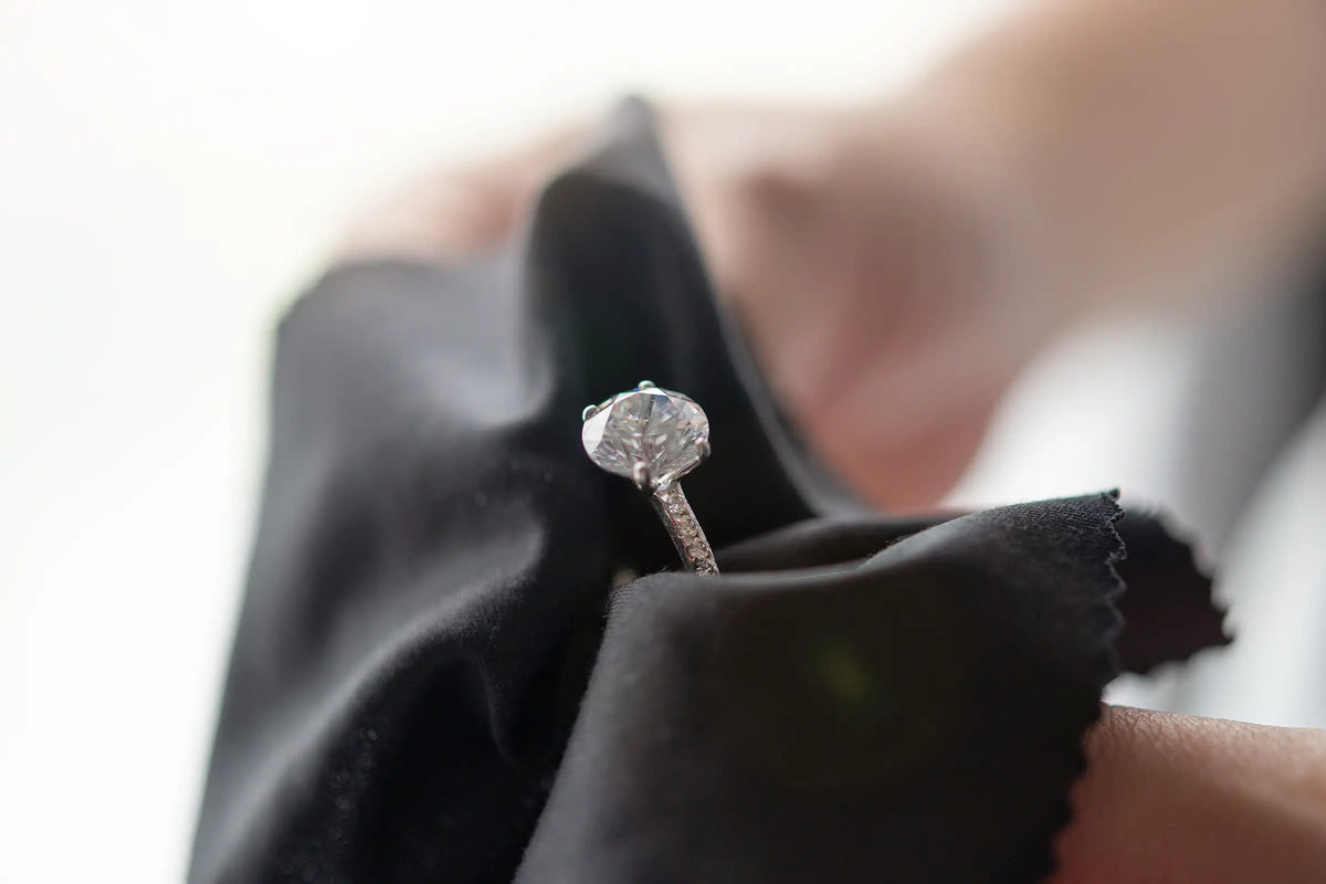 A close-up of a silver-toned diamond engagement ring held in a black cloth, with a blurred hand in the background.