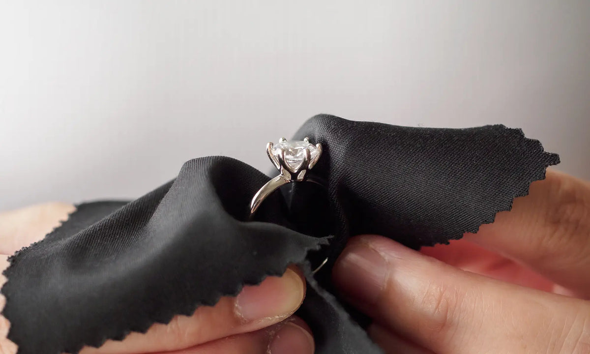 A silver-toned ring with a clear gemstone held against a black polishing cloth by hands.