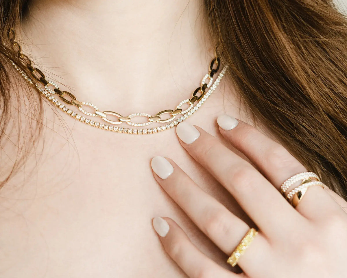 Close-up of a person wearing layered necklaces, including a gold chain and a delicate silver chain with small clear stones, along with matching rings on their hand.
