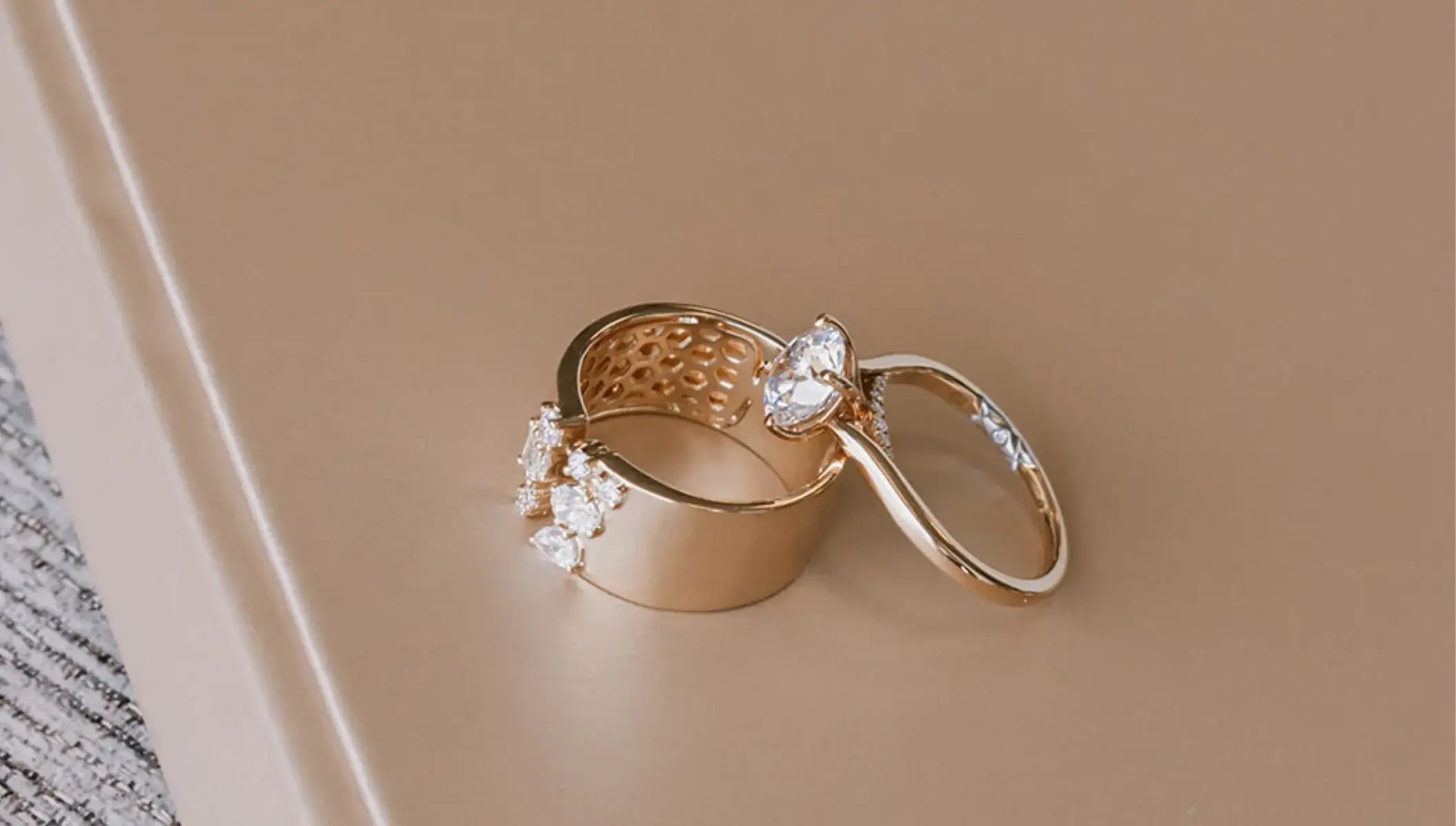 Two gold rings with clear gemstones, one featuring a textured band and the other a smooth band with a heart-shaped stone, displayed on a beige surface.