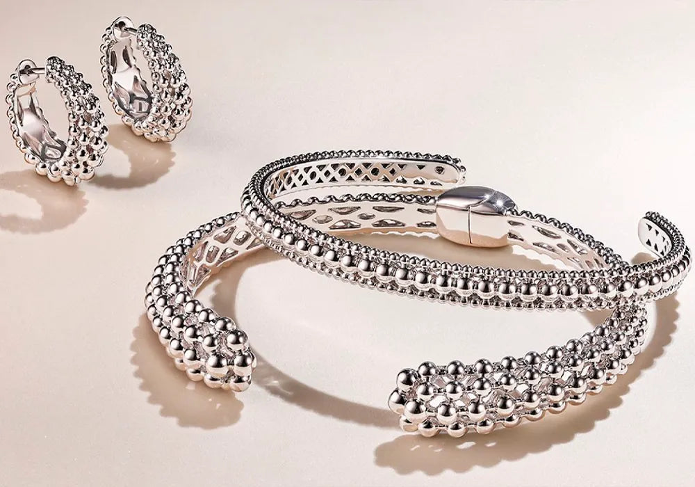 A silver-toned jewelry set featuring a textured beaded cuff bracelet, matching hoop earrings, and a slim bangle, arranged on a light beige background.