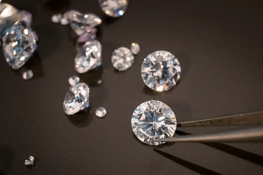 A close-up of several loose round brilliant-cut diamonds scattered on a dark reflective surface, with one diamond held by metal tweezers in the lower right.