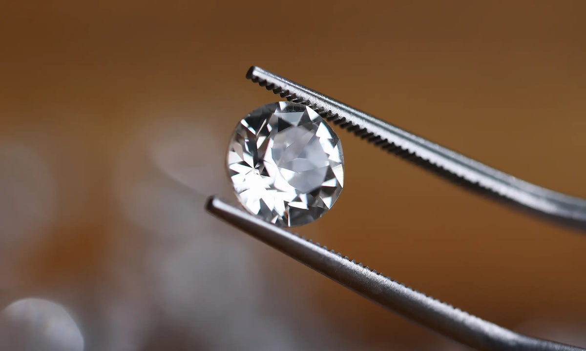 A round brilliant-cut clear gemstone held between silver metal tweezers against a warm blurred brown background.