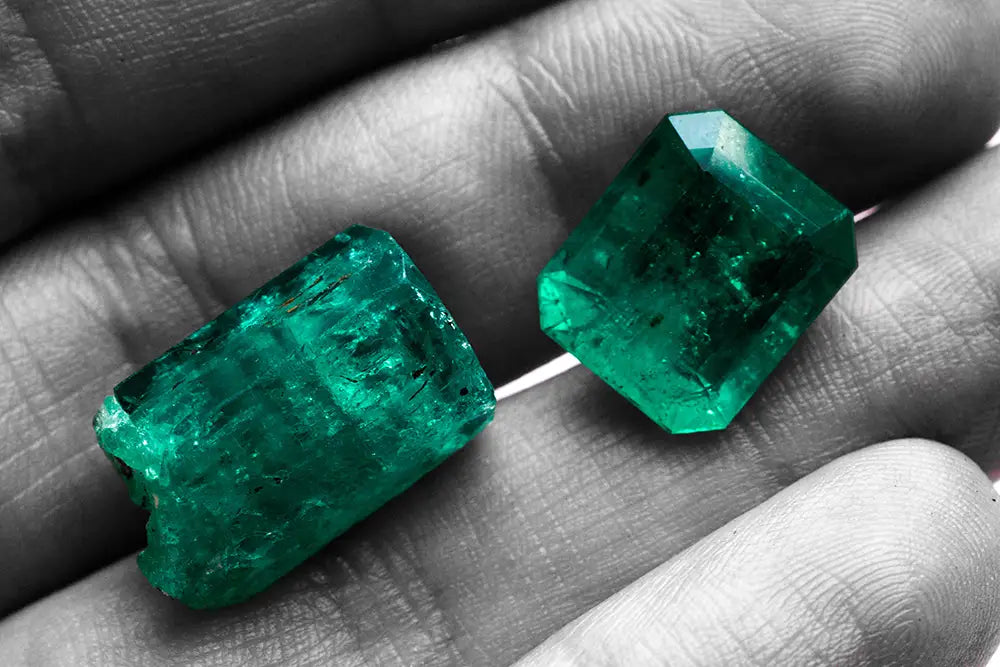 Two rough emerald-cut green gemstones resting on a person's hand, shown in color against a grayscale background of fingers and palms.