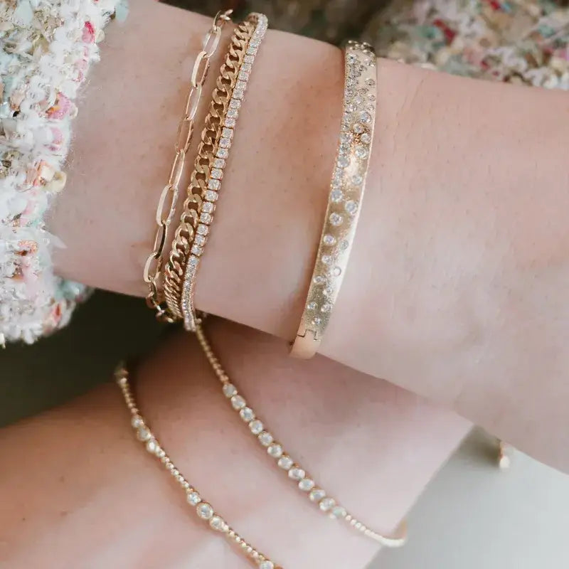 A close-up of a person's wrist and forearm wearing multiple gold-toned bracelets, including stacked chain styles and a bangle covered with clear rhinestones, with a matching rhinestone chain draped on the forearm.