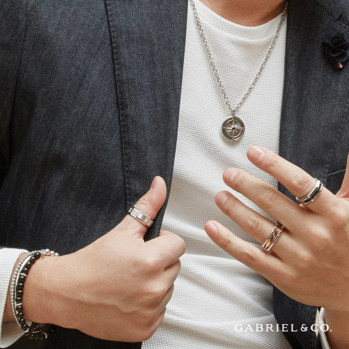 A close-up lifestyle shot of a person wearing silver jewelry, including a compass pendant necklace, multiple rings, and bracelets, styled with a white t-shirt and dark blazer. The brand name 'GABRIEL & CO.