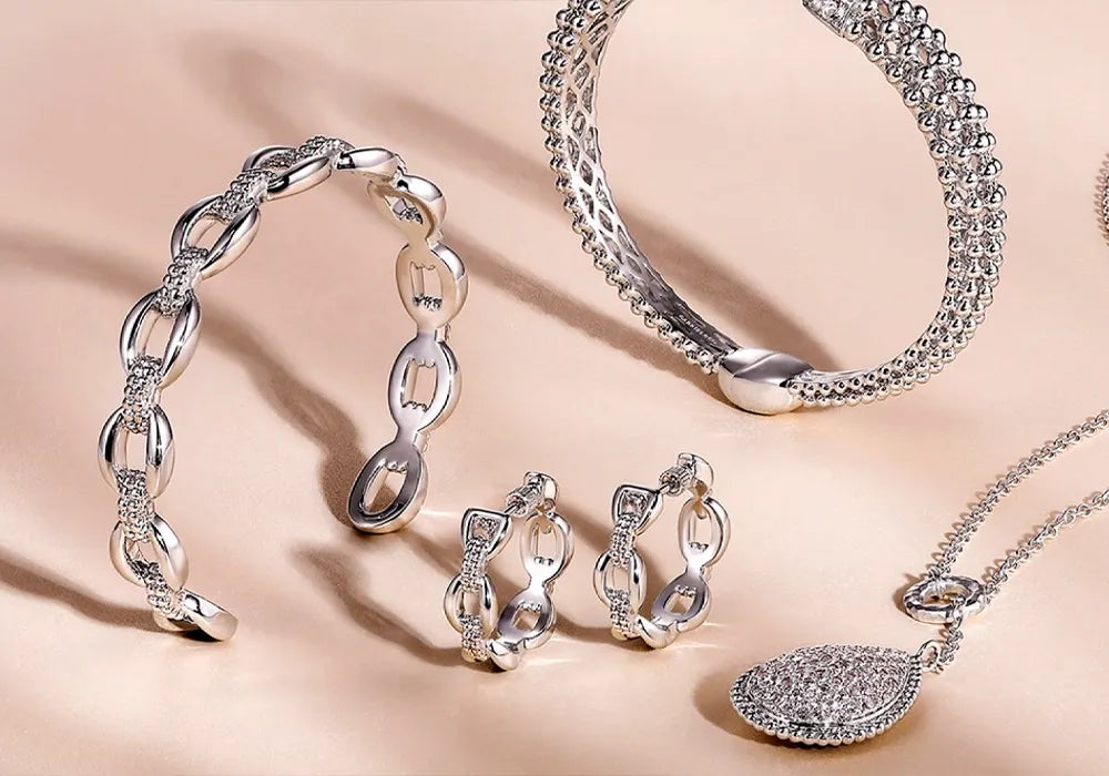 A jewelry set displayed on a warm beige background, featuring a silver-tone chain bracelet, a beaded bangle, matching earrings, a necklace with a pavé-encrusted teardrop pendant, and a textured chain necklace.