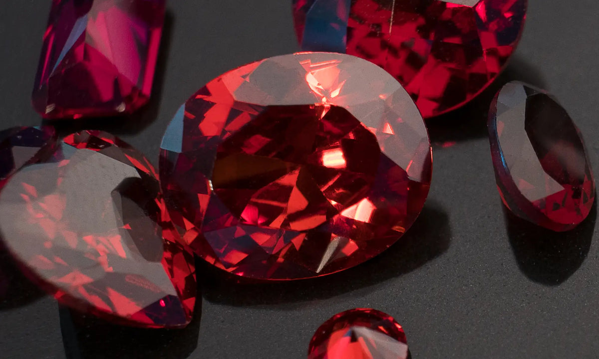 Faceted red gemstones in various shapes arranged on a dark surface, with a large oval-cut stone in the center and several smaller red stones around it.
