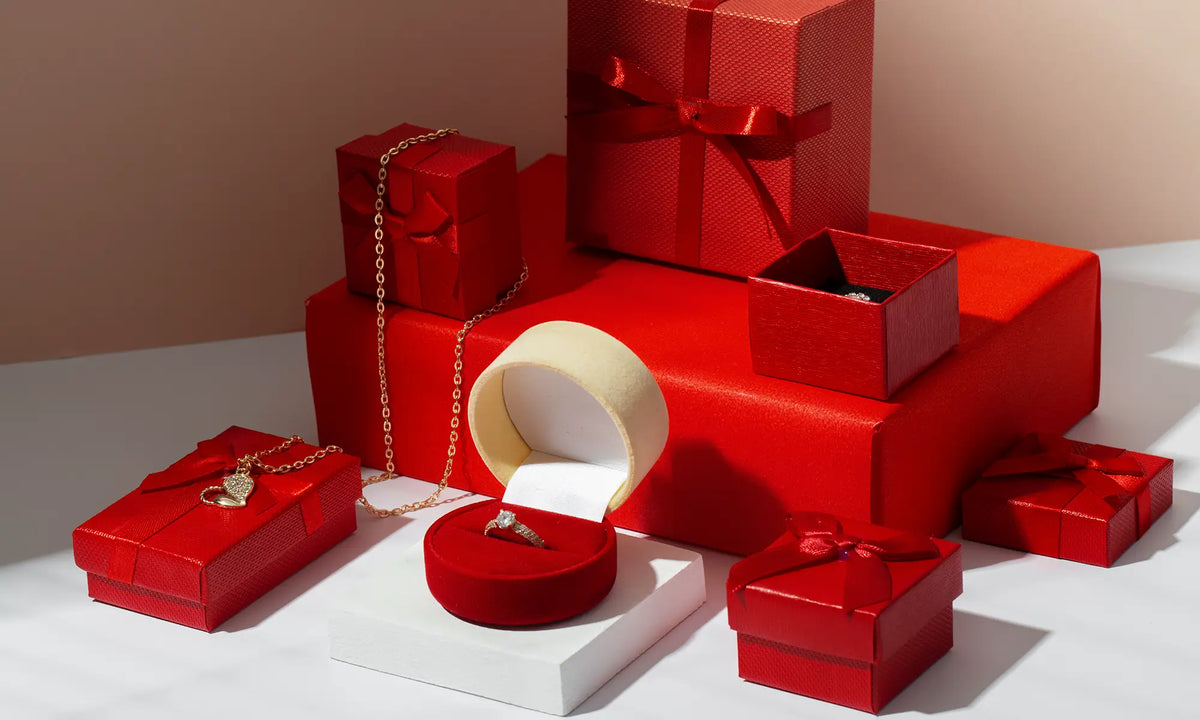 A styled arrangement of red gift boxes and jewelry boxes on a white surface against a warm beige background. An open cream-colored box and a red ring box are visible, along with a gold chain and a ring displayed on a white pedestal.