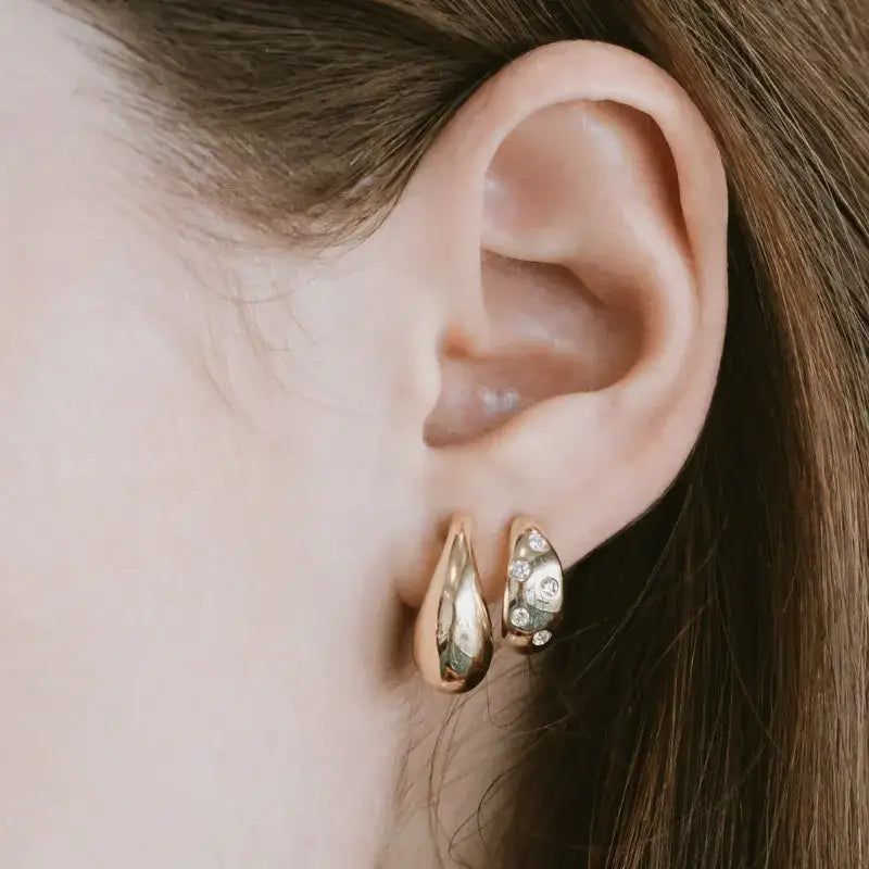 A close-up of a person's ear wearing a pair of gold hoop earrings, one plain and one decorated with small clear rhinestones.
