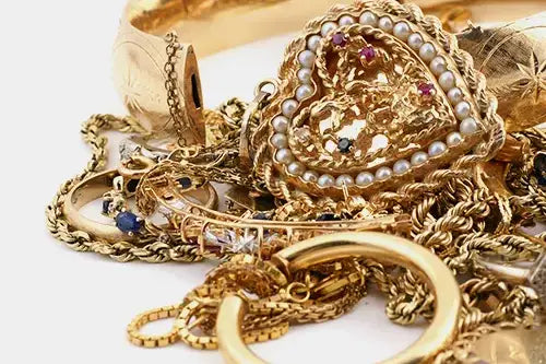 A pile of gold-toned jewelry on a white background, including chains, rings, a heart-shaped brooch with pearl-like beads and small colored stones, and a feathered accessory.