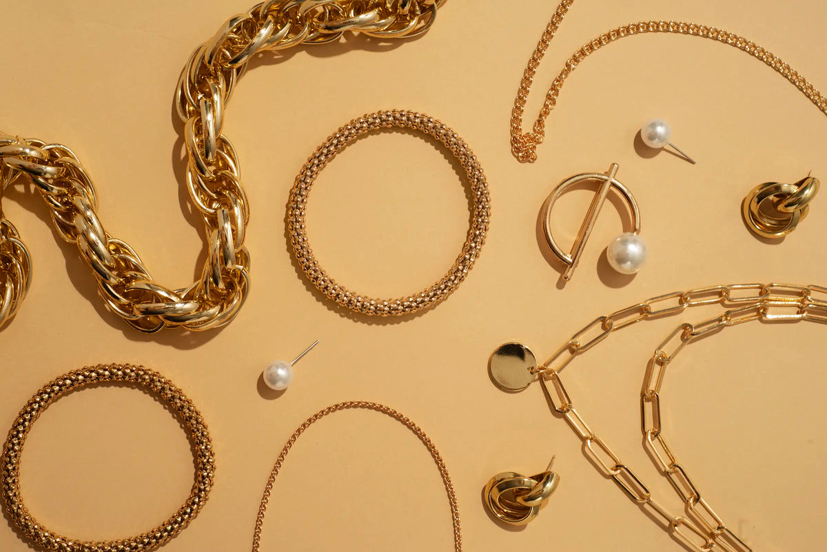 An assortment of gold-toned jewelry pieces, including chunky and chain link necklaces, beaded bracelets, stud earrings, and a pearl drop, arranged in a flat-lay pattern on a warm beige background.