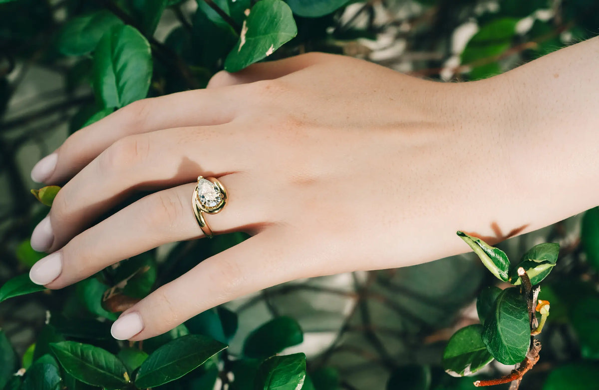 A gold ring with a teardrop-shaped clear gemstone worn on a person's ring finger, shown against a background of green leaves.