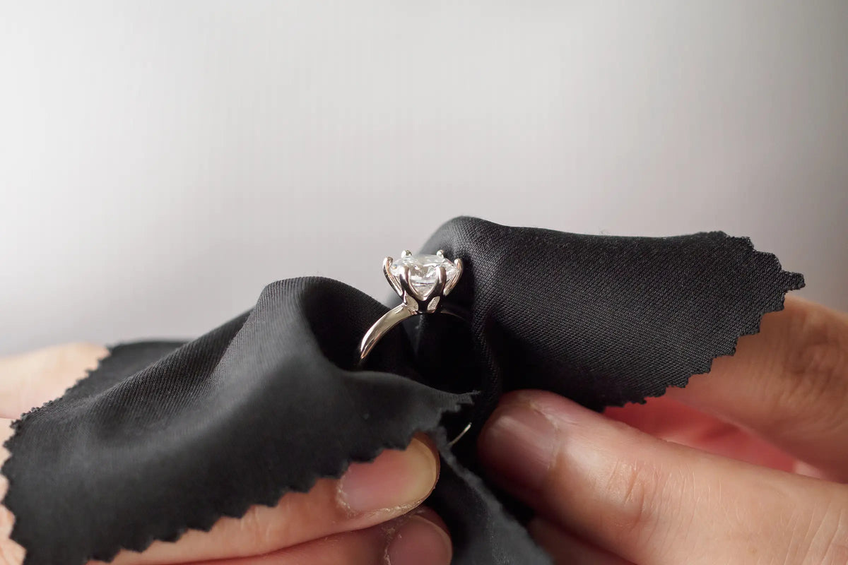 A silver-toned diamond ring held between fingers and wrapped in a black polishing cloth against a plain light gray background.