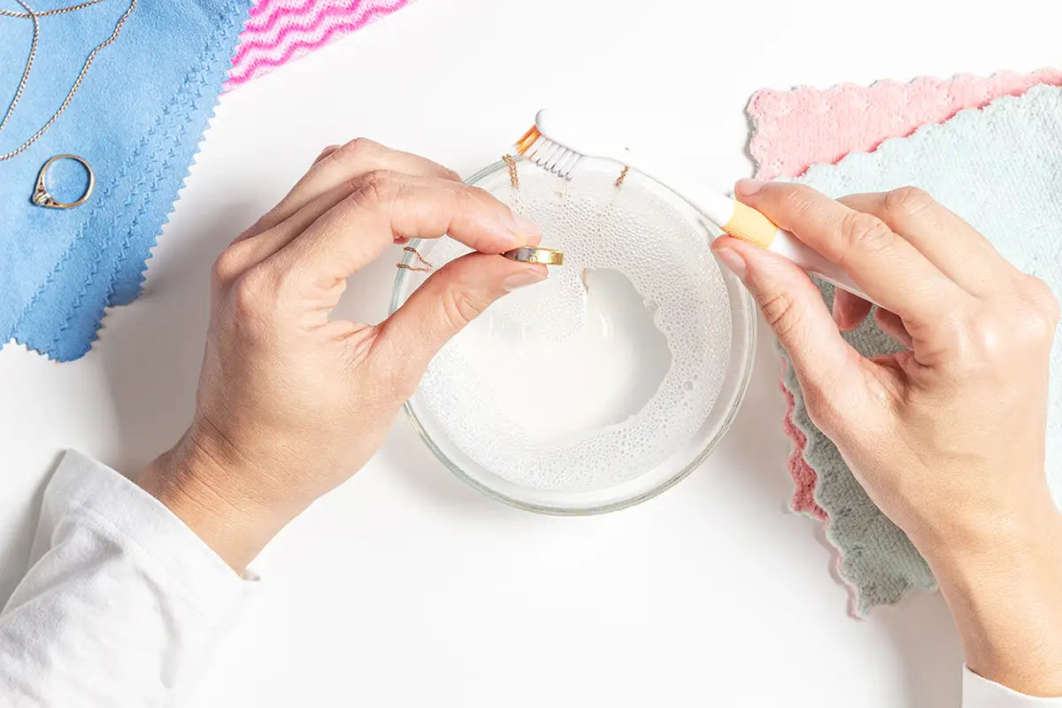 Top-down view of a person's hands using a ring warmer tool to melt a gold ring over a small glass dish, with colorful fabric cloths in the background.