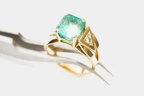A gold ring with a large cushion-cut green emerald gemstone and a small diamond accent in a triangular setting, held in a metal tweezer against a light gray background.
