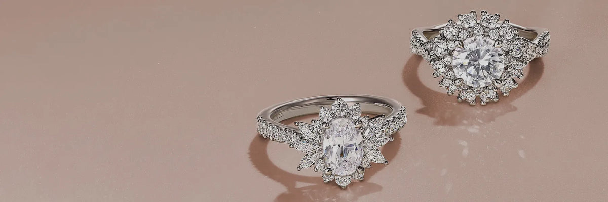 Two silver-toned diamond rings with oval center stones and halo settings, displayed on a soft pink reflective surface.