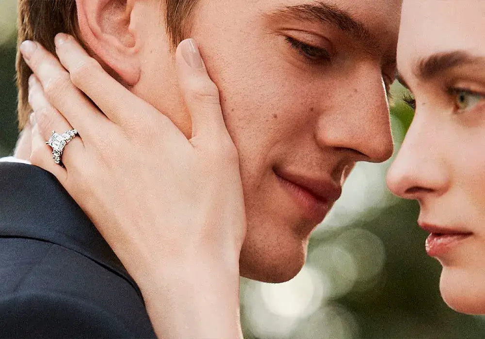 A close-up lifestyle shot of a couple with a woman's hand gently touching the man's face, wearing a diamond engagement ring on her finger.