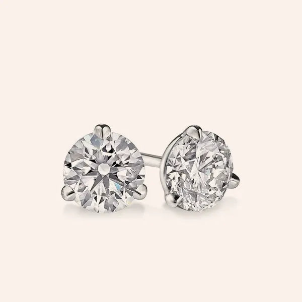 A pair of round-cut diamond stud earrings in a silver-toned setting, displayed on a clean, solid, pale pinkish-beige background.