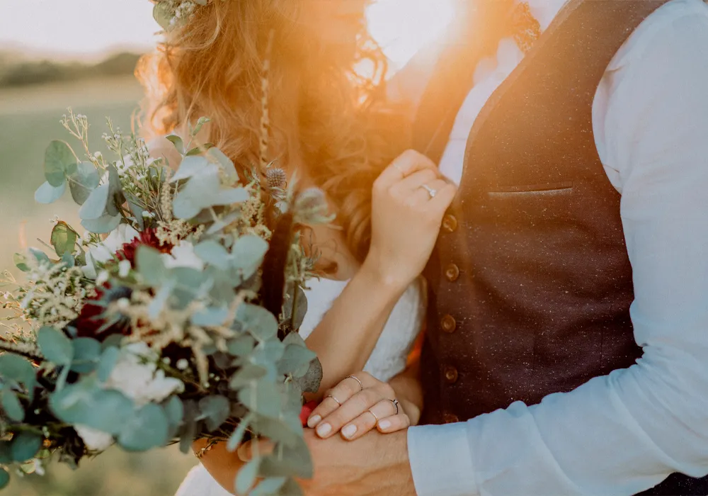 A close-up outdoor wedding photo showing a bride holding a bouquet of eucalyptus and burgundy flowers, standing next to a groom in a brown vest and white shirt, with warm sunset light and lens flare.