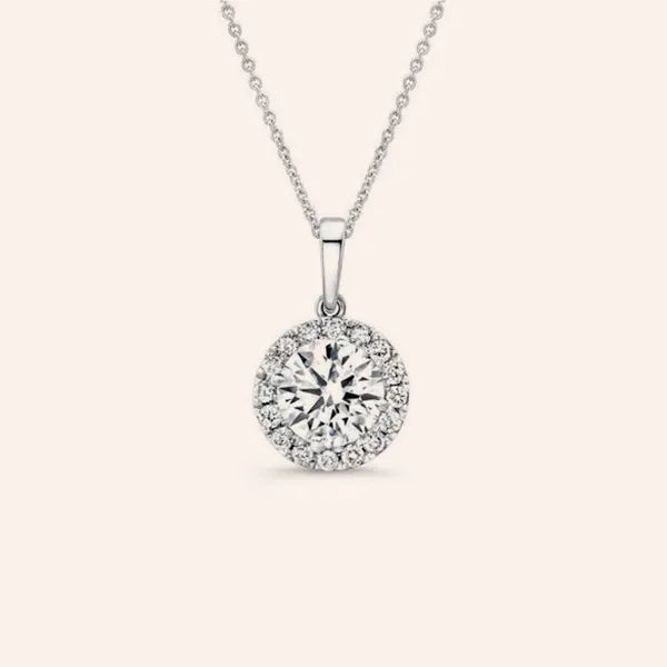 A silver pendant necklace featuring a round brilliant-cut center stone surrounded by a halo of smaller stones, suspended from a delicate silver chain against a light cream background.