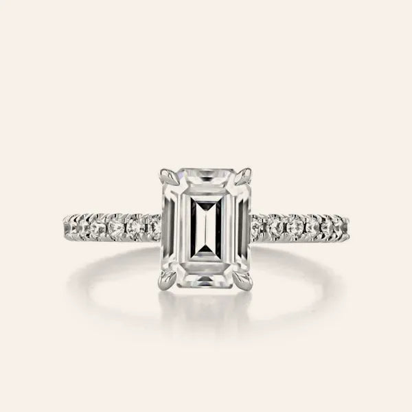 An emerald-cut diamond engagement ring with a silver-toned pavé band, centered on a light cream background.
