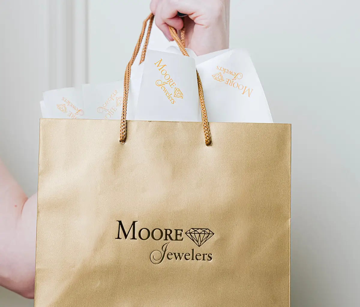 A gold paper shopping bag with twisted gold rope handles, printed with the Moore Jewelers logo, held by a hand against a light neutral background.