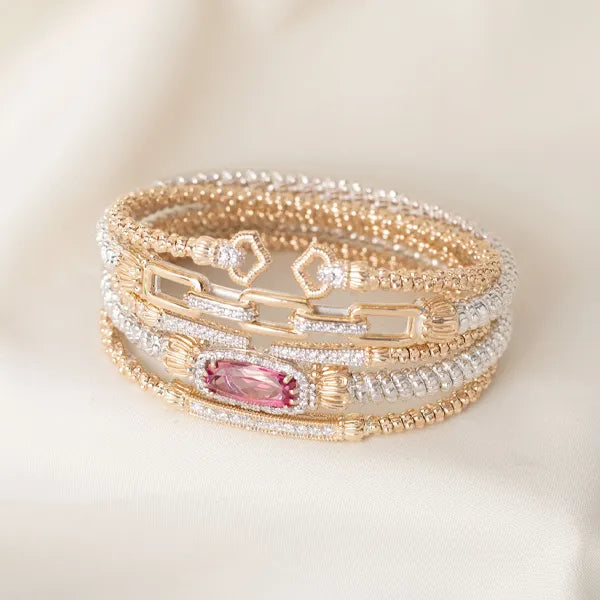 A stack of gold and silver beaded bracelets with a central pink marquise-cut gemstone, displayed on a soft cream fabric background.