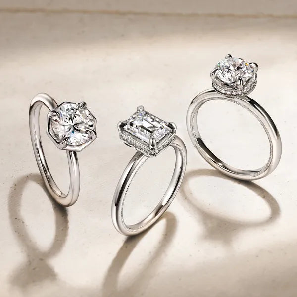 Three silver-toned rings with clear gemstones are displayed on a light beige surface. The left ring has a round brilliant-cut stone, the center ring has an emerald-cut stone, and the right ring has a large round stone surrounded by smaller stones.