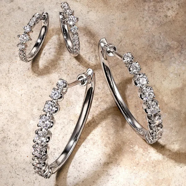 A set of silver hoop earrings with clear pavé-set stones, arranged on a textured beige stone surface.
