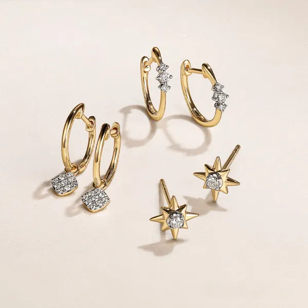 A set of gold-toned jewelry pieces on a warm cream background, including two huggie hoop earrings with small clear crystal clusters, two matching hoop earrings with larger crystal clusters, and two star-shaped stud earrings with clear crystal centers.