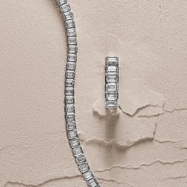 A silver-toned tennis bracelet with rectangular baguette-cut clear stones, displayed alongside a matching rectangular ring on a textured beige surface.