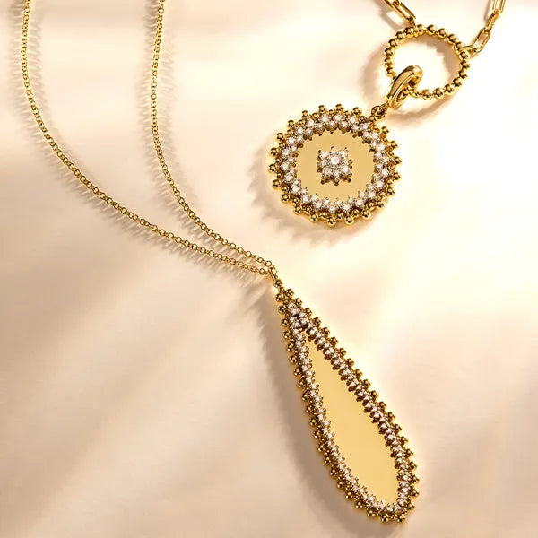 Gold-tone necklace with a teardrop-shaped pendant and a round medallion pendant, both adorned with clear rhinestones and bordered with small pearl-like accents, displayed on a soft cream fabric background.