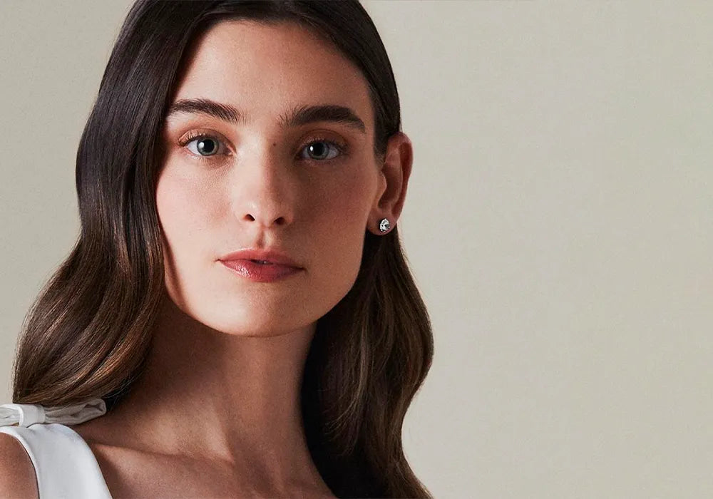 A close-up portrait of a woman with long dark brown hair wearing a small silver stud earring, against a plain light beige background.