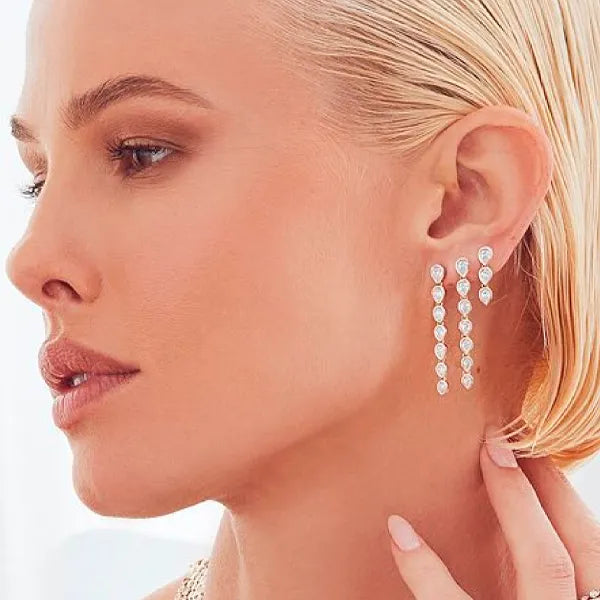 A close-up side profile of a blonde woman wearing a long, multi-drop dangle earring with sparkling clear gemstones on her ear, against a bright white background.