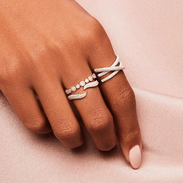 A close-up of a hand wearing two silver-toned wrap rings adorned with small clear stones, set against a soft pink satin fabric background.