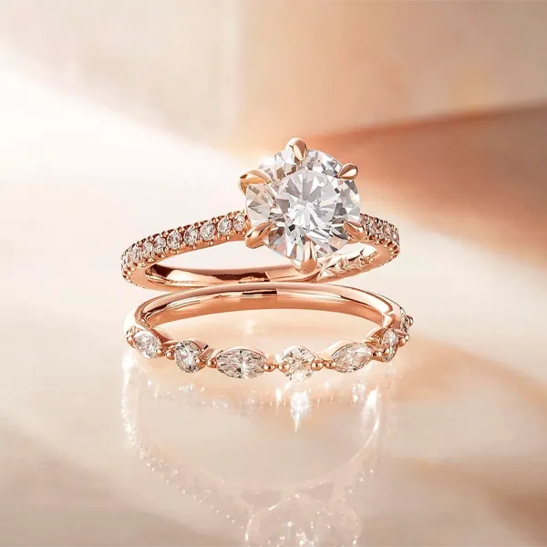 Rose gold wedding ring set featuring an engagement ring with a round brilliant-cut diamond and a matching marquise-cut diamond band, displayed on a warm beige reflective surface.