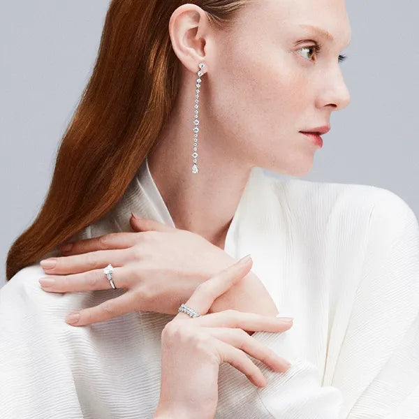 A close-up of a woman in a white textured top wearing a long silver drop earring and matching silver rings on her fingers, shown in profile against a light gray background.