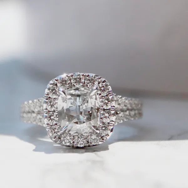 A radiant cut diamond halo ring in silver-colored metal, featuring a prominent cushion-cut center diamond surrounded by a halo and a pavé-set band, resting on a light marble surface.